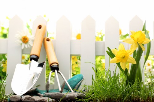 The image depicts a section of a residential garden with a focus on gardening tools and planting elements. In the foreground, there are two small hand tools, a trowel and a hand fork, resting on the ground. Next to the tools, there is a pair of gardening gloves made of fabric, also lying on the soil. To the right, vibrant yellow daffodils with green leaves are growing directly from the soil, adding a splash of natural colour to the scene. Behind these flowers, there is a neatly trimmed hedge and a white picket fence, creating a charming enclosed outdoor space. The background appears to feature additional plantings and possibly a garden path, though these are not fully visible. The overall setting is illuminated by natural daylight, suggesting a bright, mild weather condition typical of well-maintained front or back gardens in Chiswick or nearby areas. The arrangement reflects careful gardening and outdoor maintenance practices available through professional services such as Garden Maintenance Chiswick, highlighting a tidy and attractive garden environment suitable for leisure or outdoor enjoyment.