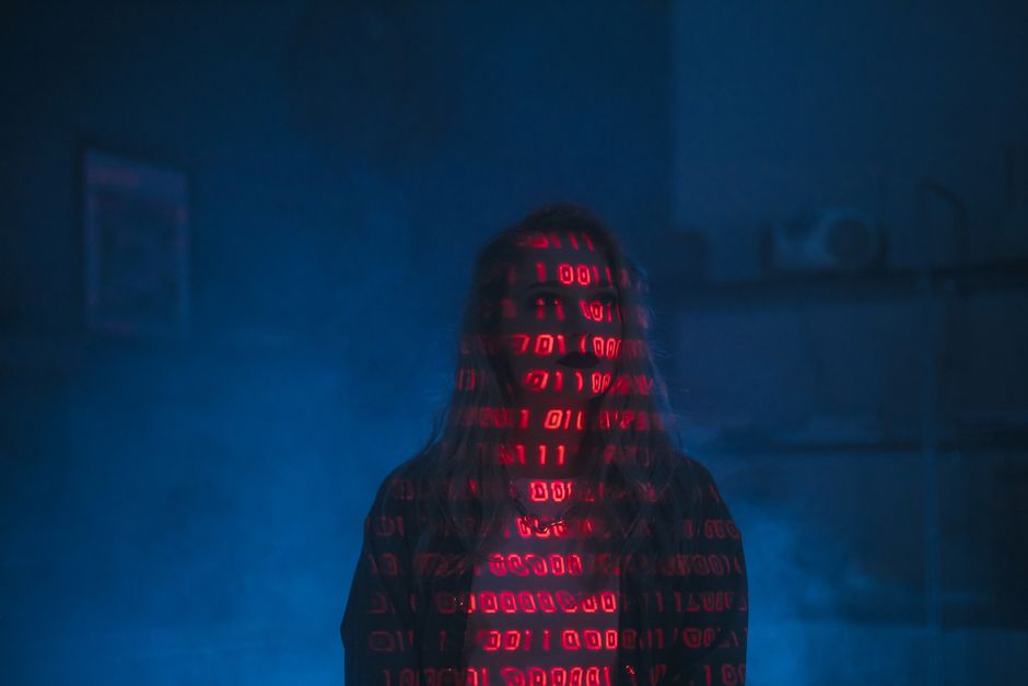 A person standing in a dimly lit room with a dark background, their face obscured by shadows. They are wearing a hoodie with a pattern of red binary code projected onto it, creating an image of cascad