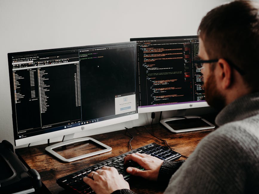 A person with short brown hair and glasses is seated at a wooden desk, working on a computer with dual monitors. The left monitor displays a file explorer window and a login prompt, while the right mo