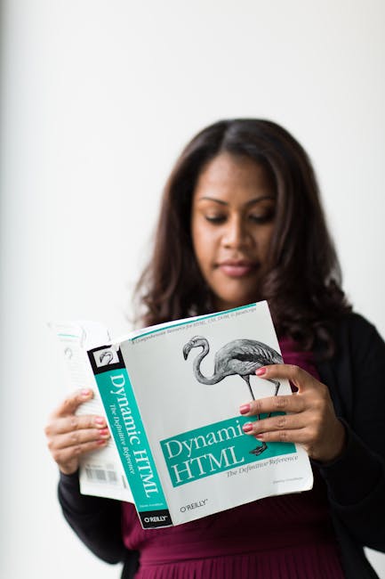 The image shows a woman with dark, shoulder-length hair reading a book titled 'Dynamic HTML,' which features a black and white illustration of a flamingo on the cover. She is wearing a dark blazer ove